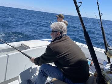 Two anglers actively fishing on a boat in open water, with one reeling in a line while the other observes the ocean.