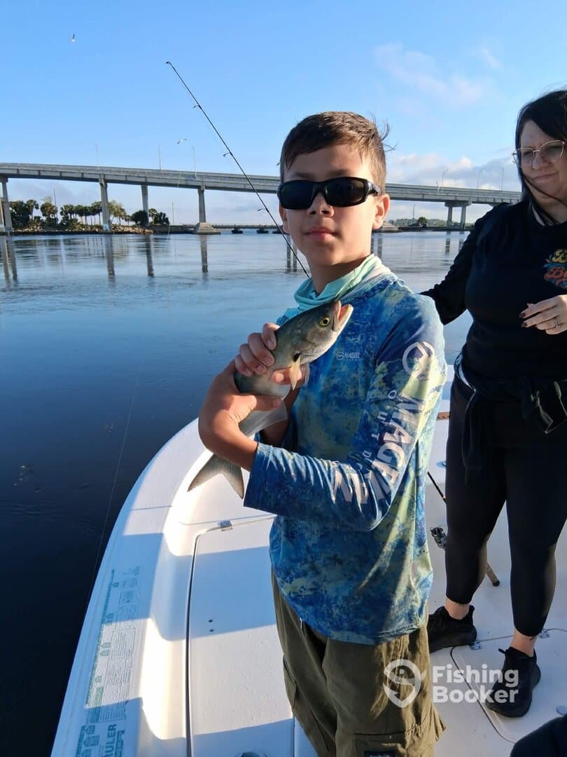 A young angler proudly holding a fish while fishing from a boat, with a scenic bridge in the background.