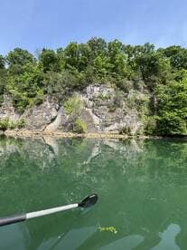 A scenic view of a calm lake surrounded by lush greenery and rocky cliffs, with a paddle partially visible in the foreground.