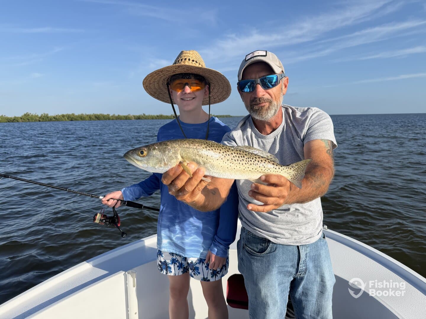 A young angler and an adult proudly displaying a large Speckled Trout while fishing on a boat in a coastal waterway.