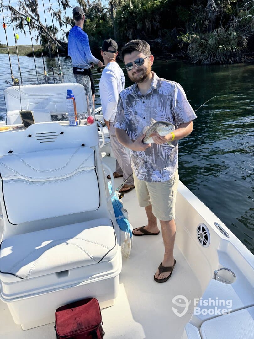 A proud angler holding a freshly caught fish on a boat, surrounded by fellow fishermen in a scenic waterway.