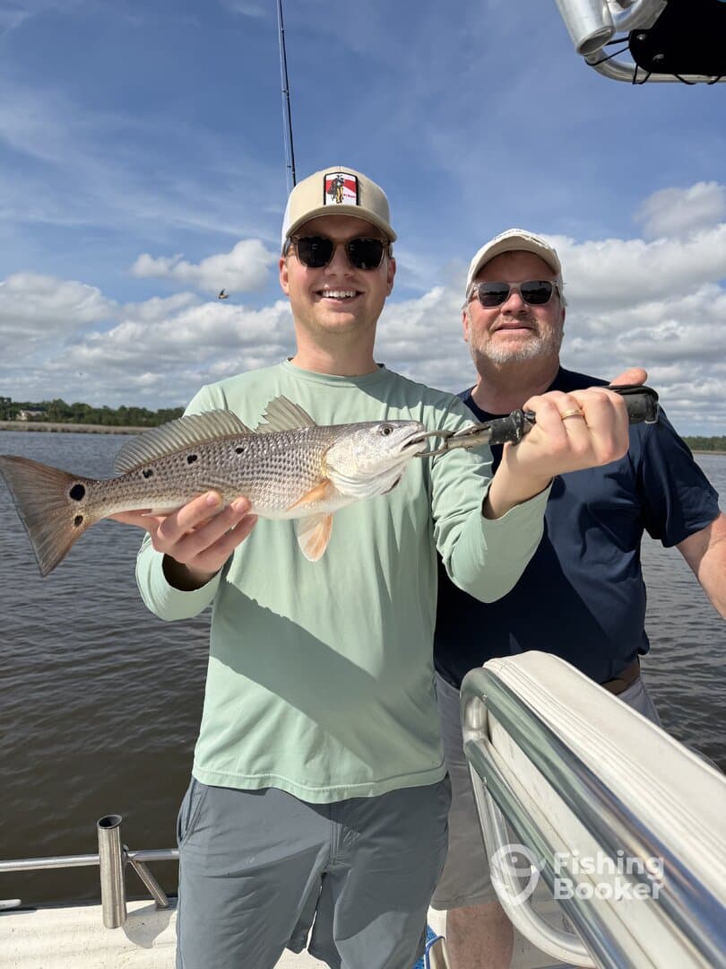 Two anglers proudly displaying a Redfish on a boat during a sunny fishing trip.