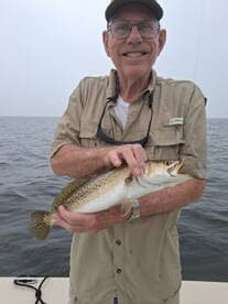 An elderly angler proudly displaying a Speckled Trout while fishing on a calm day.