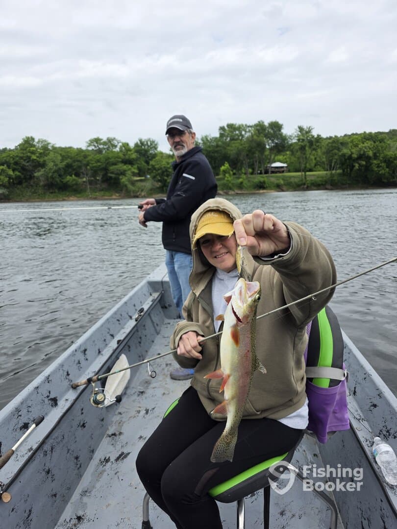 A young angler proudly displaying a freshly caught Rainbow Trout while fishing from a small boat on a calm river.