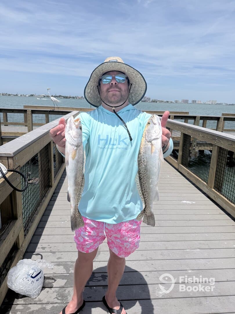 Angler proudly displaying two Speckled Trout caught from a pier, showcasing a successful day of fishing.
