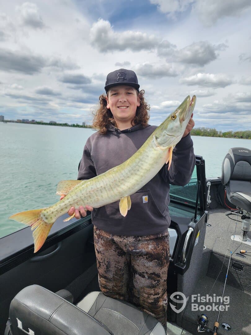 Young angler proudly holding a large Muskie while fishing from a boat on a clear day.