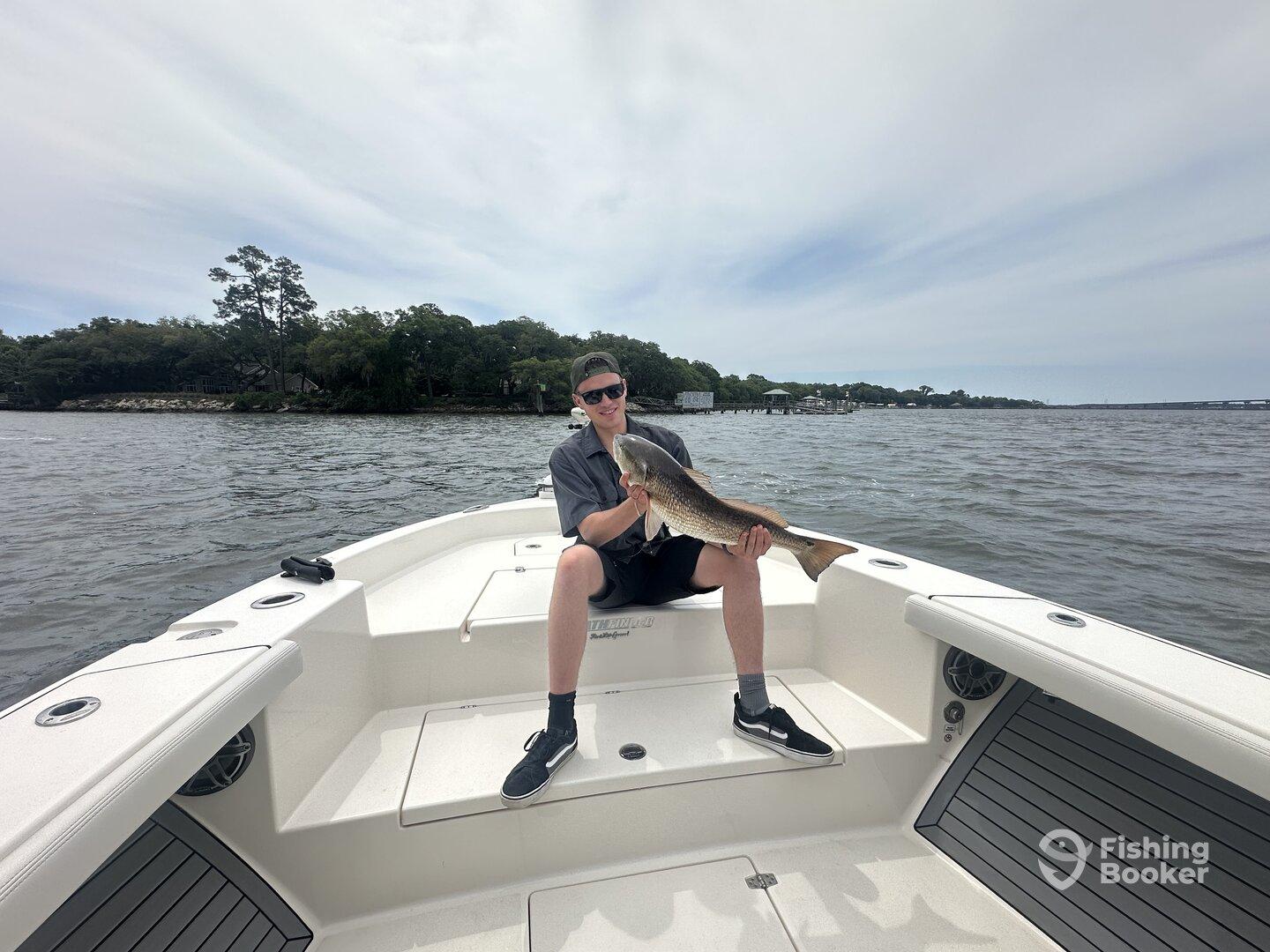 A young angler proudly displaying a large Redfish while seated on a boat in a scenic waterway.