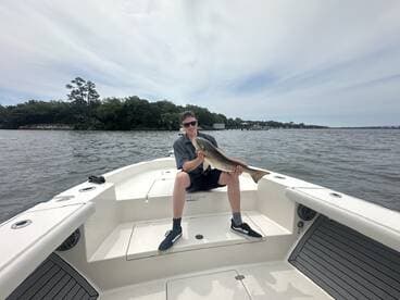 A young angler proudly displaying a large Redfish while seated on a boat in a scenic waterway.