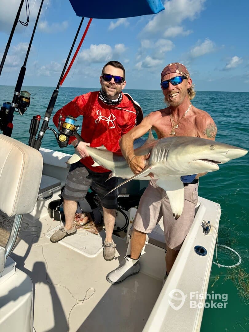 Two anglers proudly displaying a large shark caught during a fishing trip on a boat in clear waters.