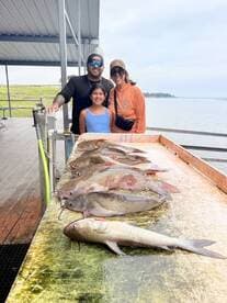 A family poses with their catch of catfish displayed on a cleaning table at a fishing dock, showcasing a successful day on the water.