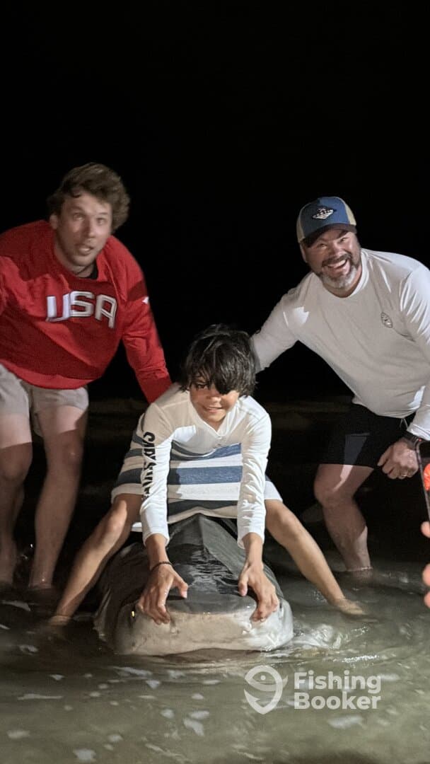 A young angler poses with a large shark while being assisted by two adults during a nighttime fishing adventure.
