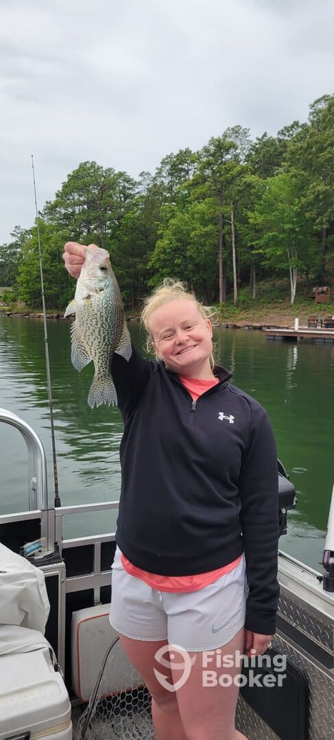 A young angler proudly displaying a large Crappie while fishing from a boat in a scenic lake setting.