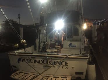 The fishing charter boat 'Pure Indulgence' is docked at night, illuminated by onboard lights, showcasing its fishing gear and equipment.