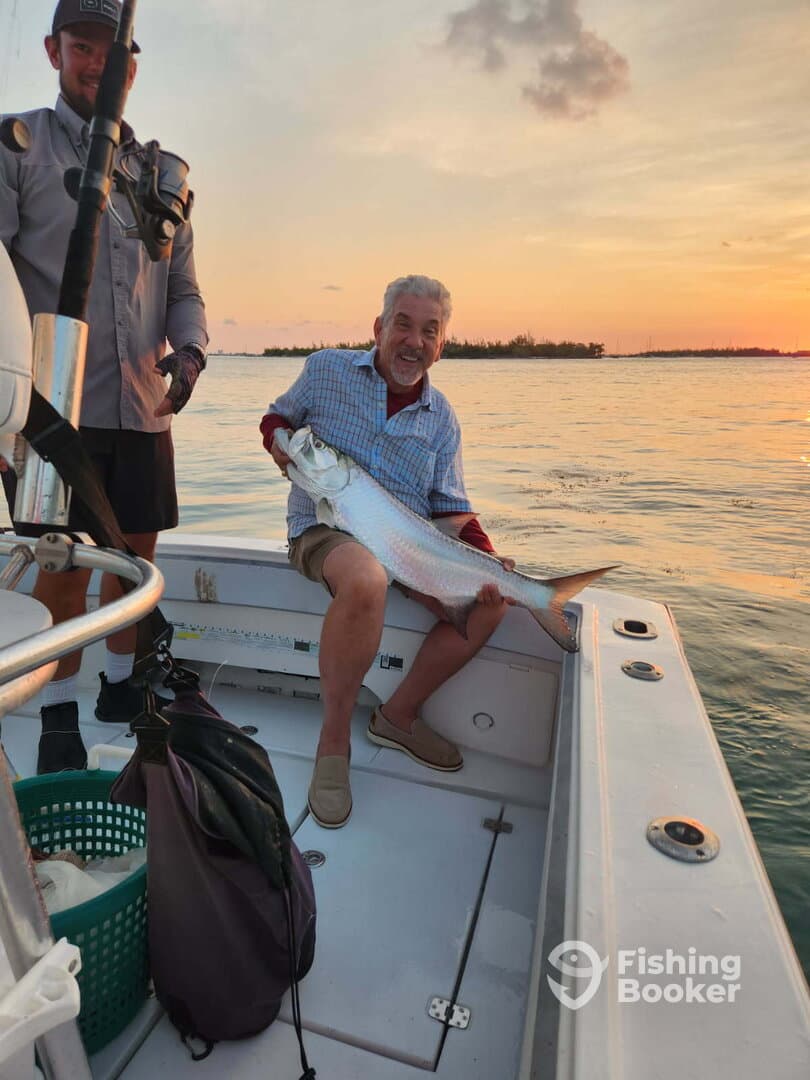 An excited angler proudly displays a large Tarpon while enjoying a sunset fishing trip on a boat.