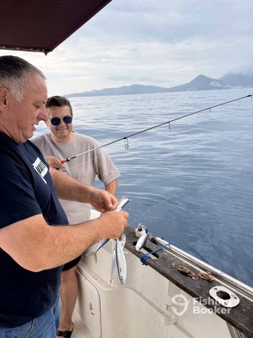 Two anglers actively fishing on a boat, preparing bait with a scenic backdrop of calm waters and distant mountains.