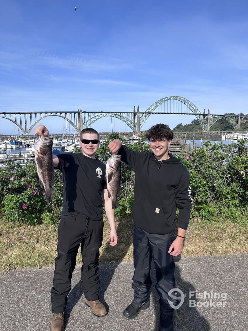 Two young anglers proudly displaying their catch of rockfish in front of a scenic marina and bridge backdrop.