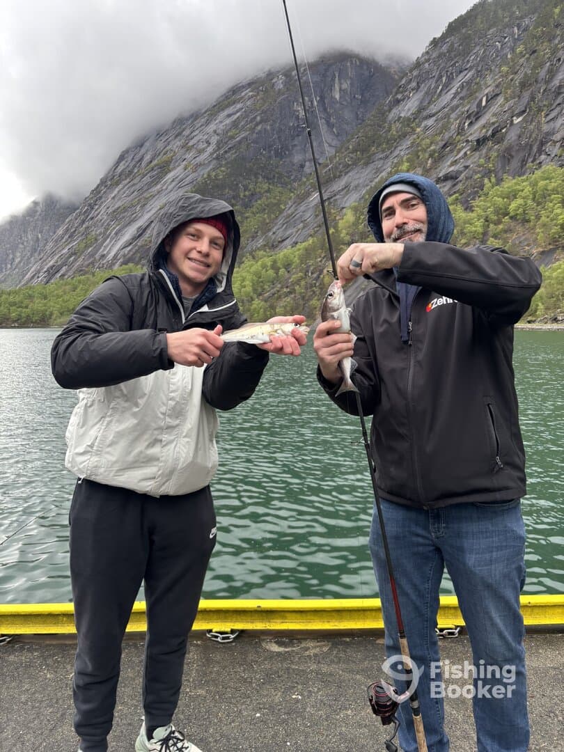 Two anglers proudly displaying their catches, a couple of fish, while fishing in a scenic location surrounded by mountains.