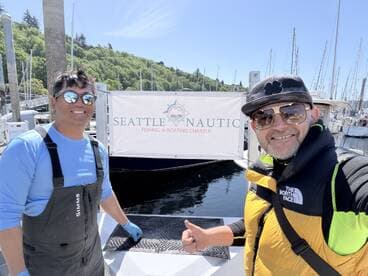 Two smiling individuals pose in front of the Seattle Nautic Fishing & Boating Charter sign, showcasing a sunny day at the marina.