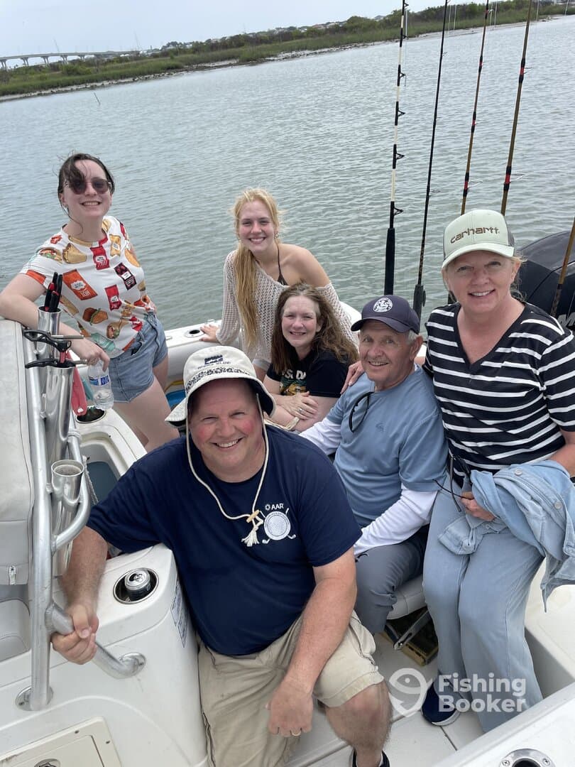 A cheerful group of five adults and teens enjoying a day on a fishing boat, with fishing rods visible in the background.