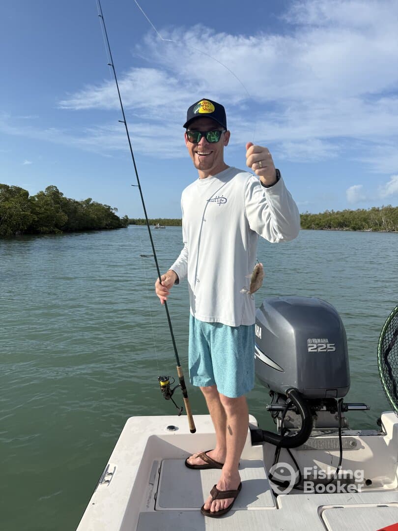 Angler smiling while reeling in a small fish on a sunny day in a mangrove area.