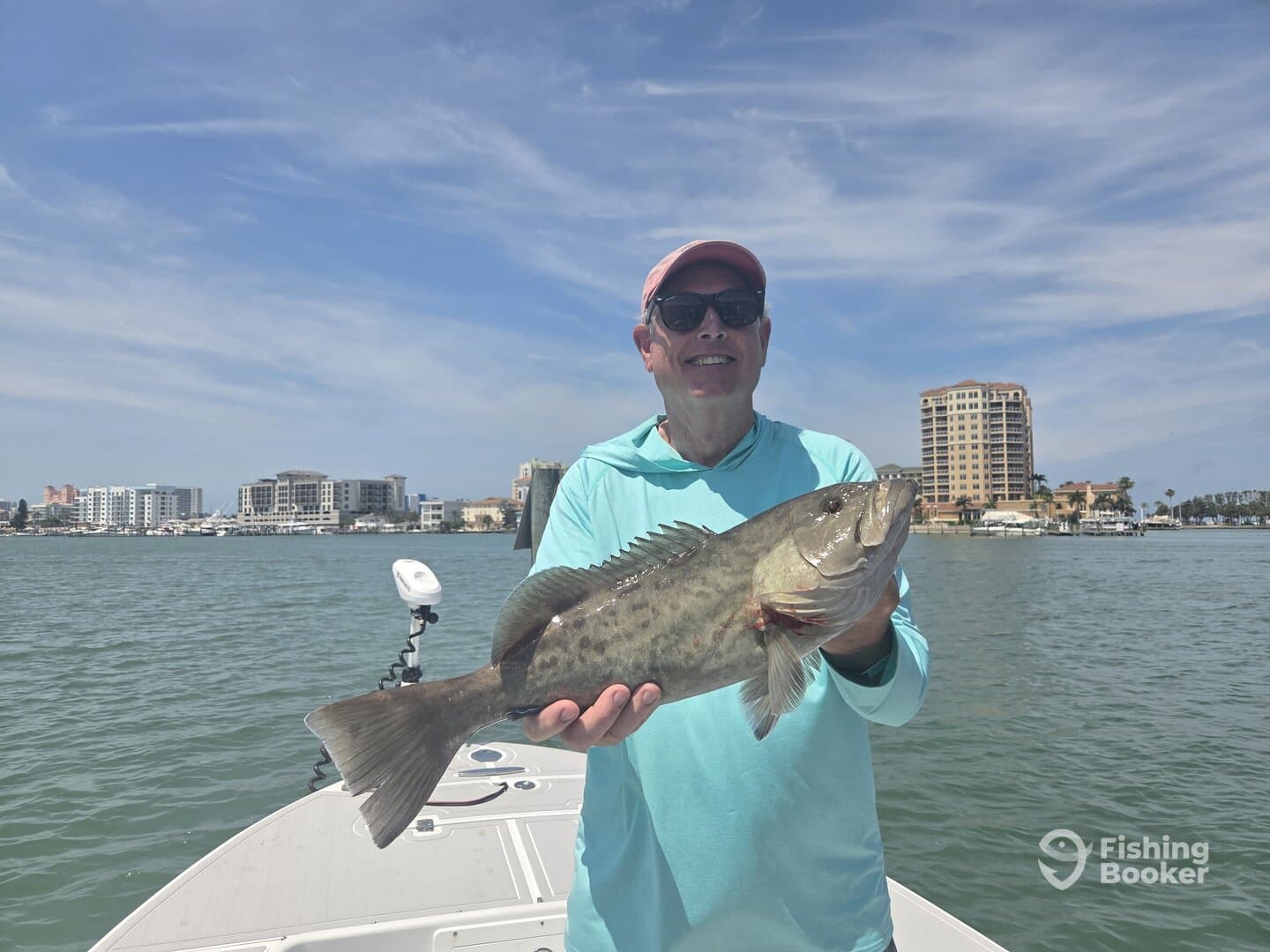 Angler proudly displaying a large Grouper while fishing in a coastal area, with a scenic backdrop of buildings and boats.