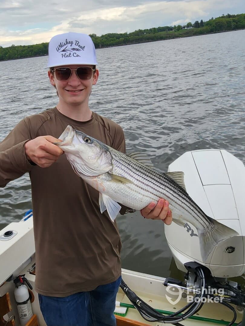 A young angler proudly displaying a large Striped Bass while fishing on a boat, showcasing a successful day on the water.