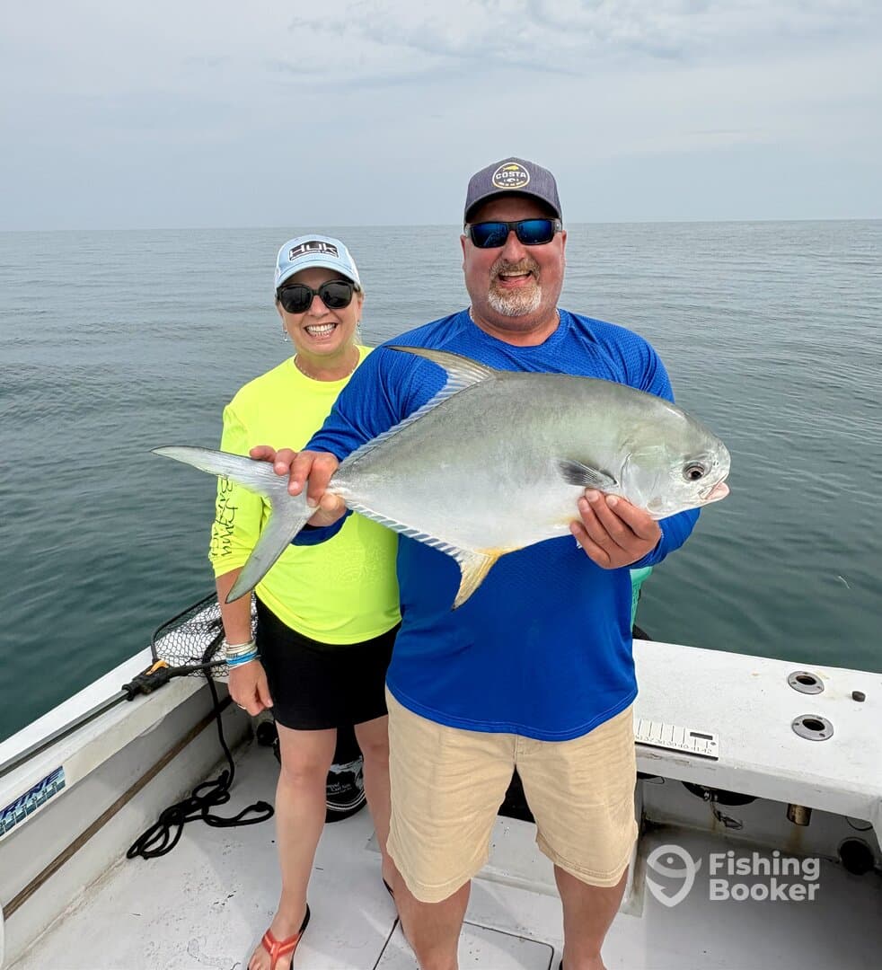 A proud couple displaying a large Amberjack while fishing on a boat in calm waters.