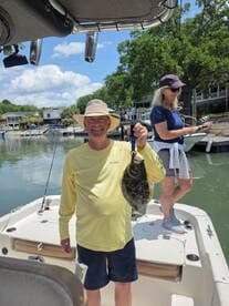 A proud angler holding a Flounder while fishing on a boat, with another person in the background preparing to cast.