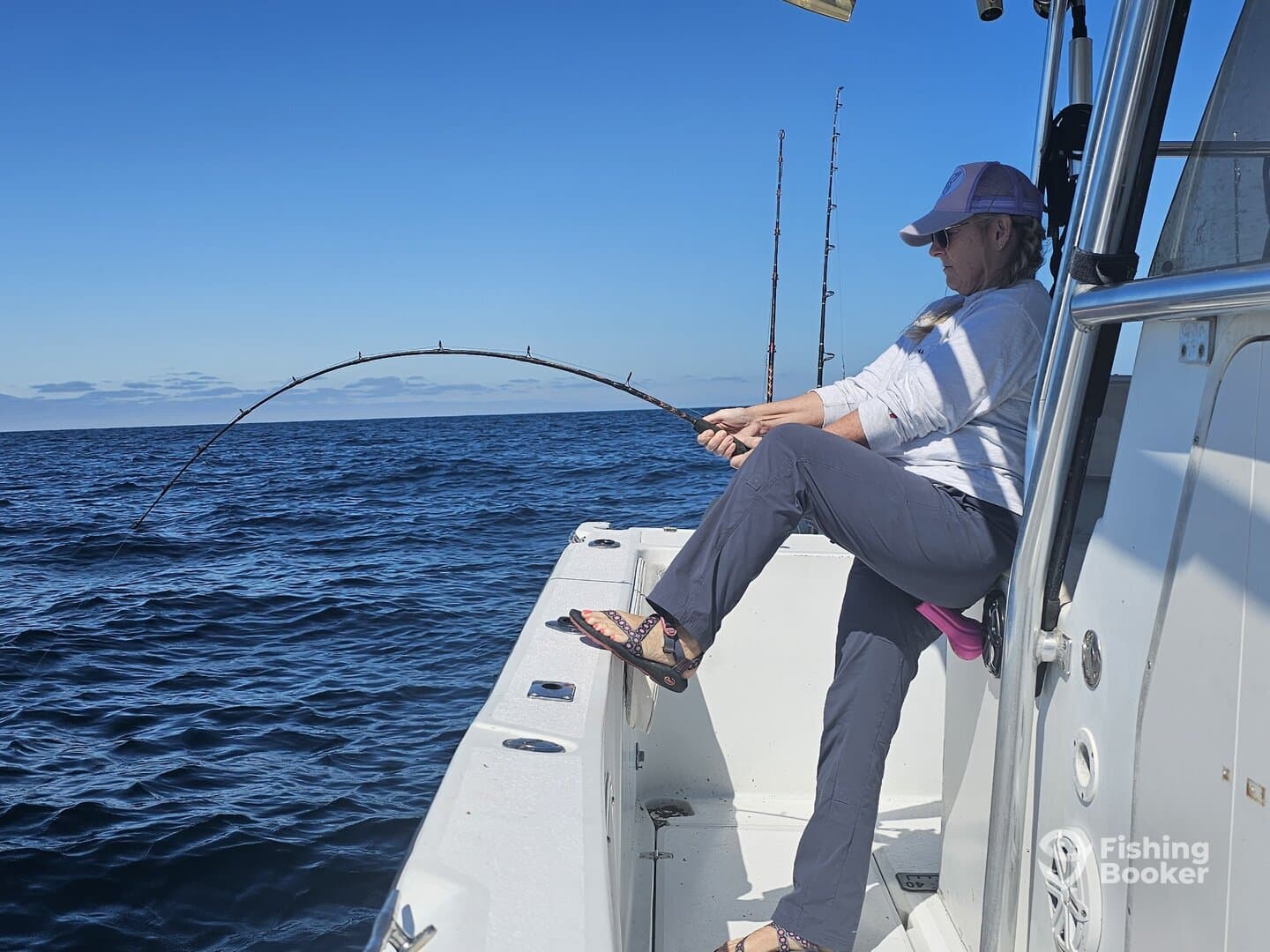 A female angler is actively reeling in a fish while seated on a boat, showcasing the excitement of deep-sea fishing in clear blue waters.
