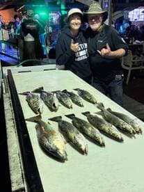 A group of anglers proudly displaying their catch of speckled trout on a board at a lively fishing venue.