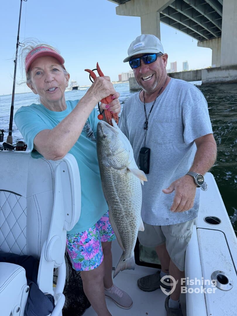 A woman proudly holds a large Black Drum while fishing with a guide under a bridge, showcasing a successful day on the water.