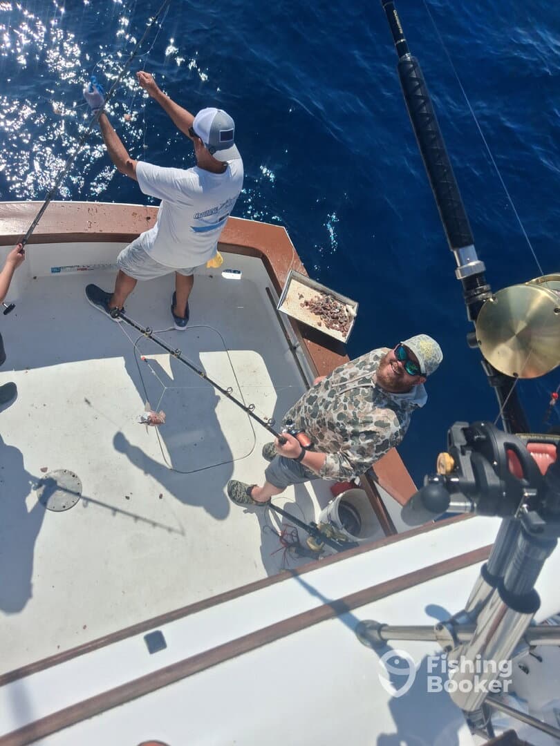Two anglers actively fishing on a charter boat, with one reeling in a catch while the other prepares to cast. The vibrant blue ocean provides a stunning backdrop.