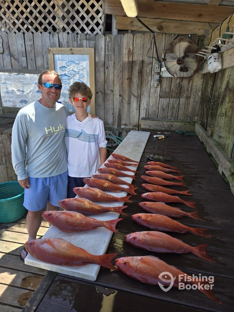 A proud father and son duo stand beside a display of freshly caught Red Snapper on a cleaning table, showcasing their successful fishing trip.