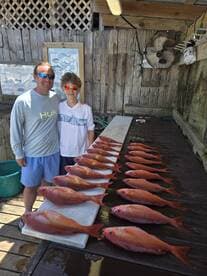 A proud father and son duo stand beside a display of freshly caught Red Snapper on a cleaning table, showcasing their successful fishing trip.