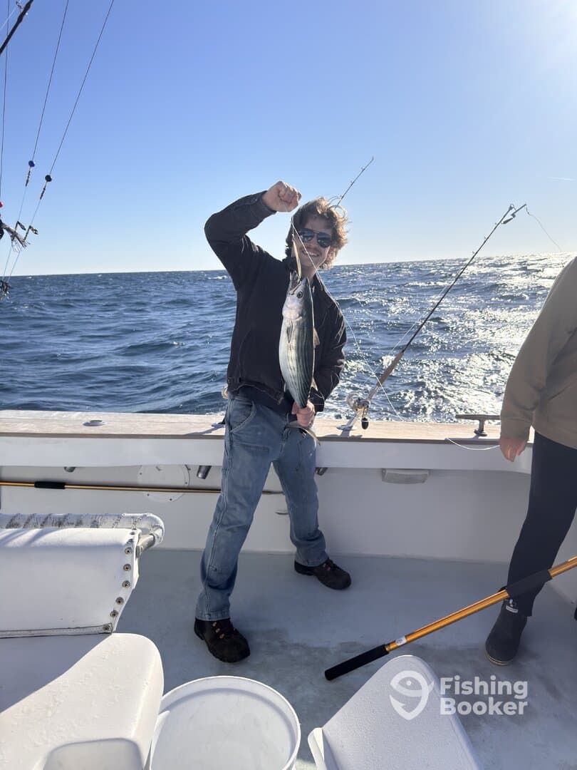 A young angler proudly displaying a striped bass while fishing on a boat, with a clear blue sky and ocean in the background.
