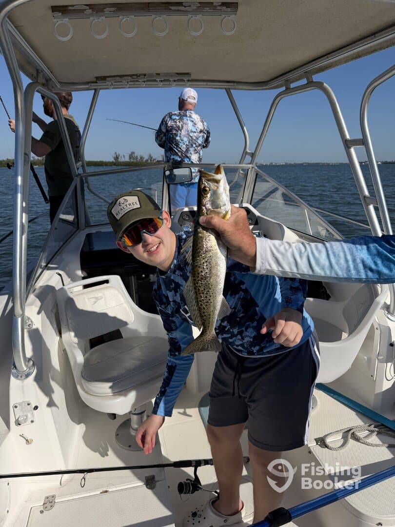 A young angler proudly displaying a Speckled Trout while fishing on a boat, with fellow anglers in the background.