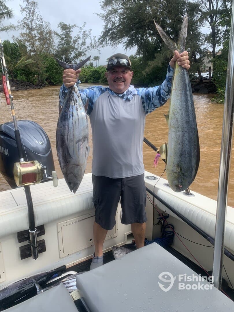 A proud angler displaying two large fish, a Mahi Mahi and a Tuna, aboard a fishing boat in a tropical setting.
