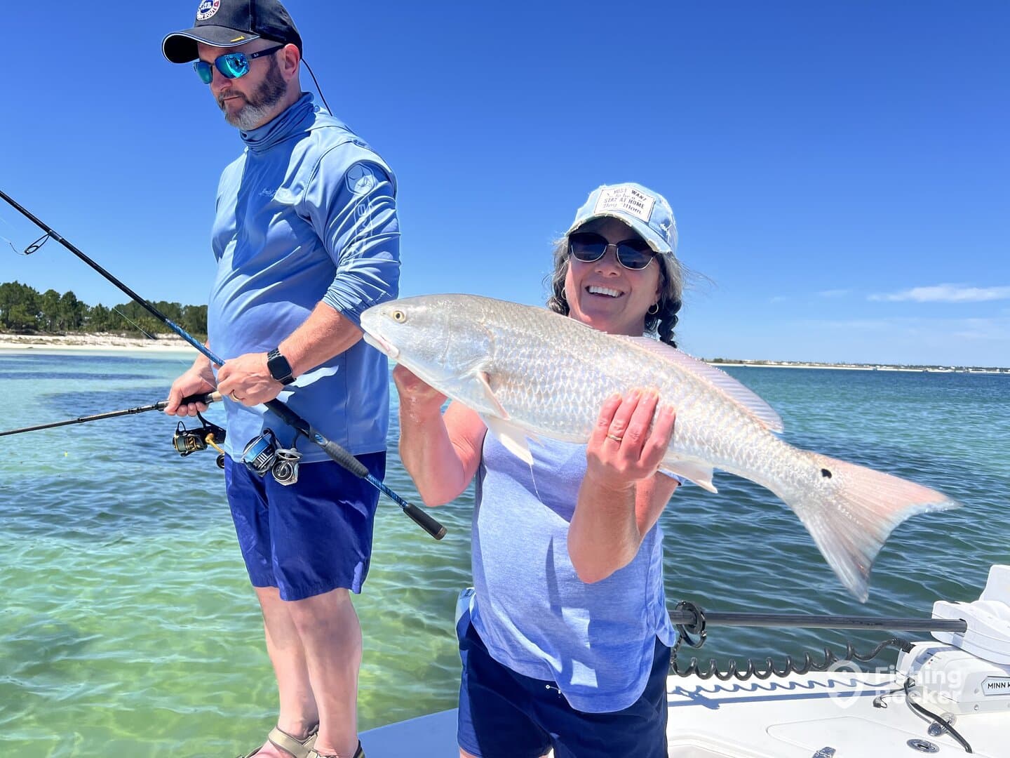 A woman proudly displays a large Redfish while fishing with a companion in a scenic coastal environment.