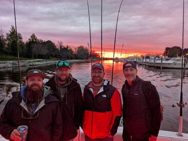Group of four anglers enjoying a sunset view from their boat, with fishing rods in the background.