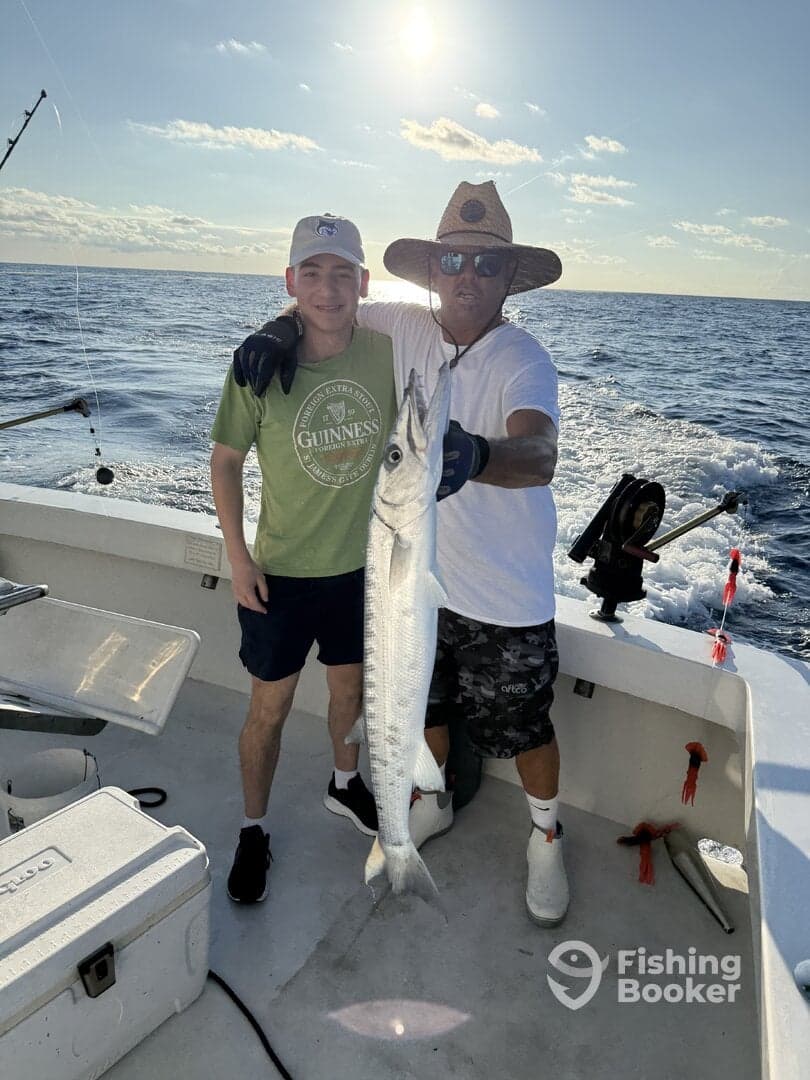 A young angler and an adult proudly display a large Barracuda on a fishing boat, enjoying a successful day on the water.