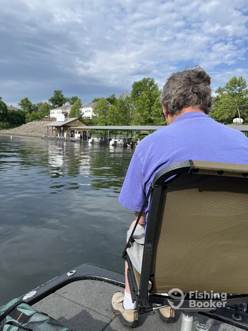 A lone angler seated in a fishing boat, enjoying a peaceful day on the water, with a scenic view of a dock and trees in the background.