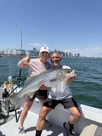 Two anglers proudly displaying a large Striped Bass while fishing in a scenic urban environment.