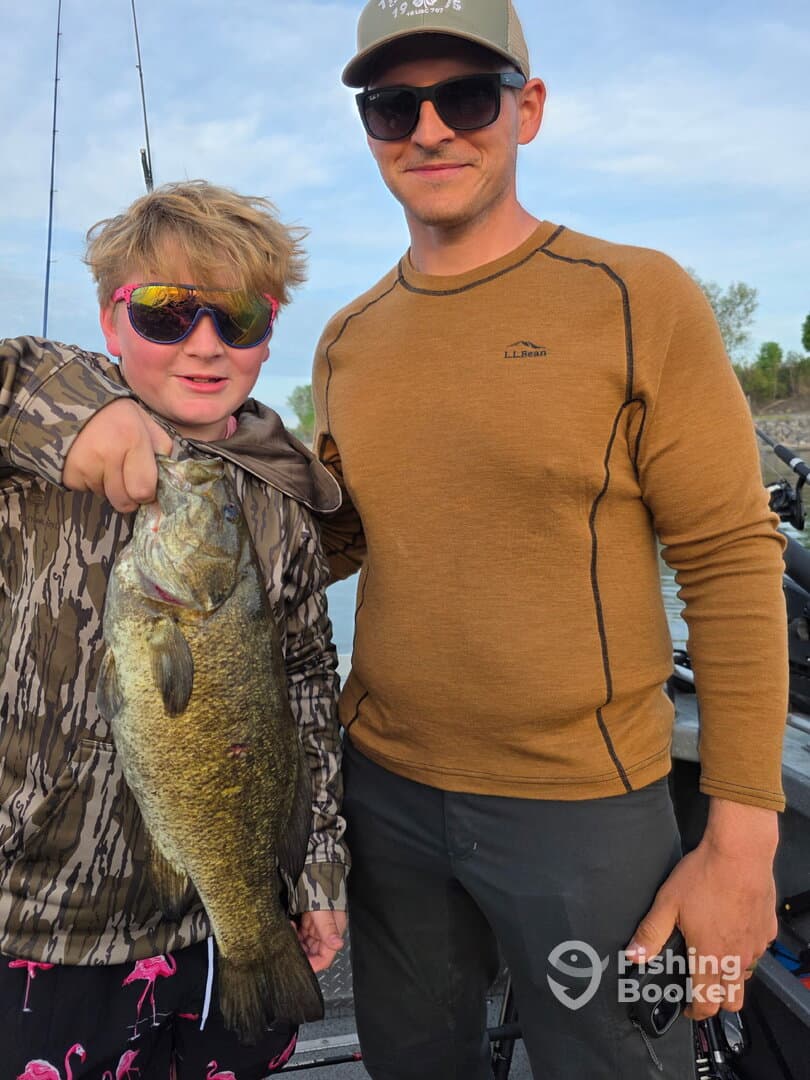 A young angler proudly displaying a large Bass while fishing with a family member on a sunny day.