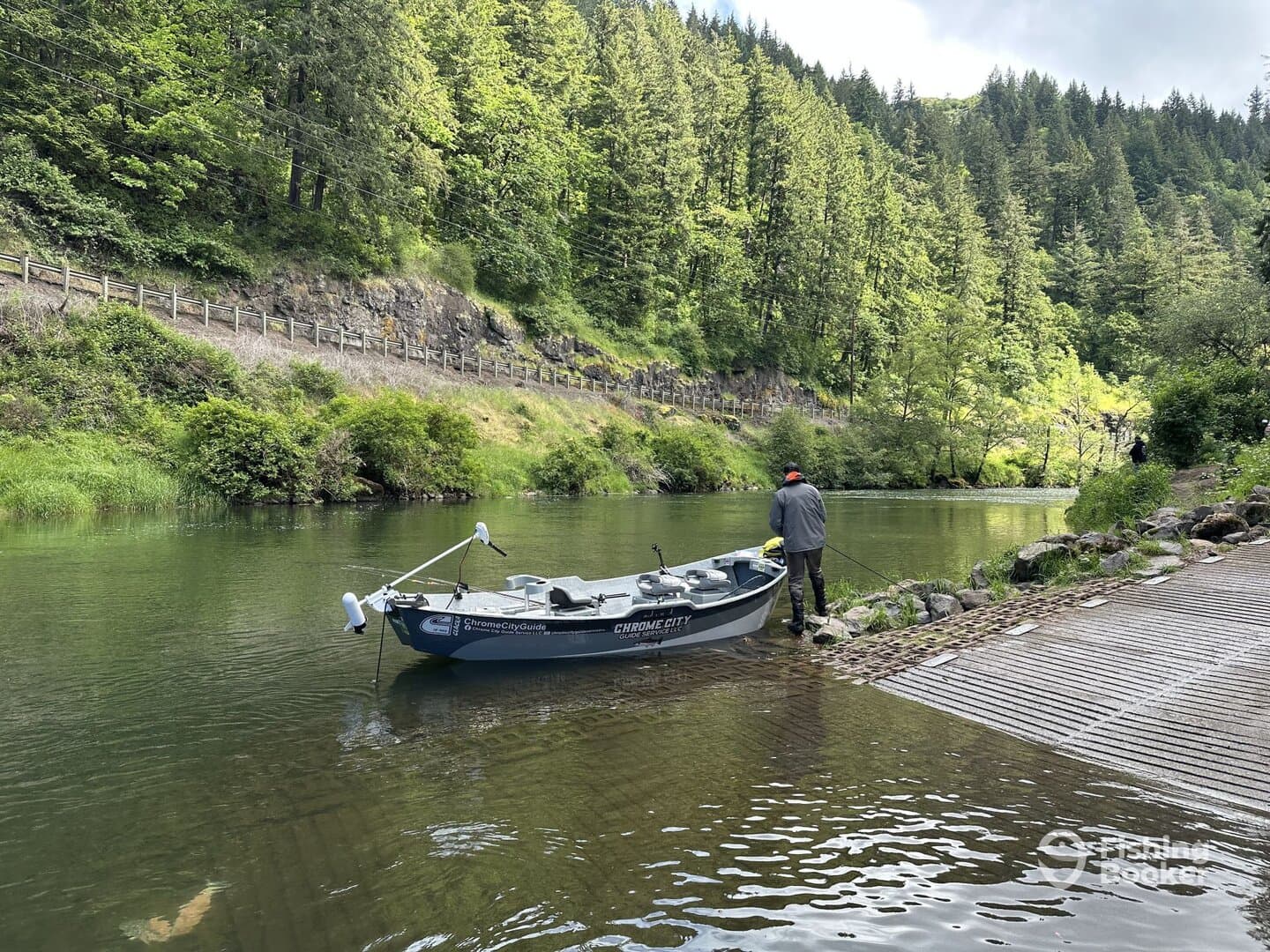 A fisherman stands by a drift boat on a serene river, surrounded by lush greenery, preparing for a day of fishing.