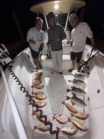 Three anglers posing on a boat with a well-arranged display of various fish, including Snapper and Grouper, after a successful night fishing trip.