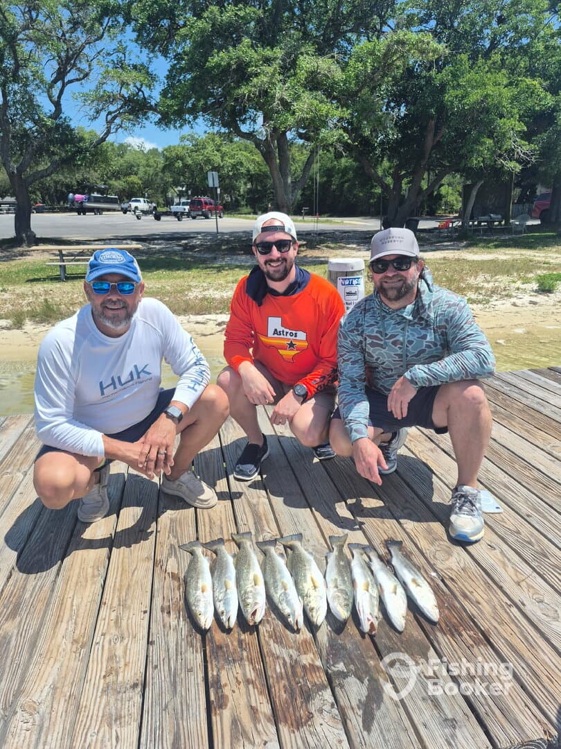 A group of three anglers proudly displaying their catch of Speckled Trout on a dock after a successful fishing trip.