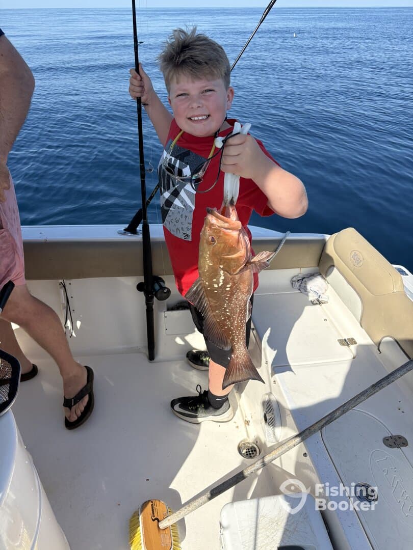 A young angler proudly displaying a large Grouper while fishing on a boat in calm waters.