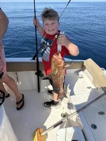 A young angler proudly displaying a large Grouper while fishing on a boat in calm waters.