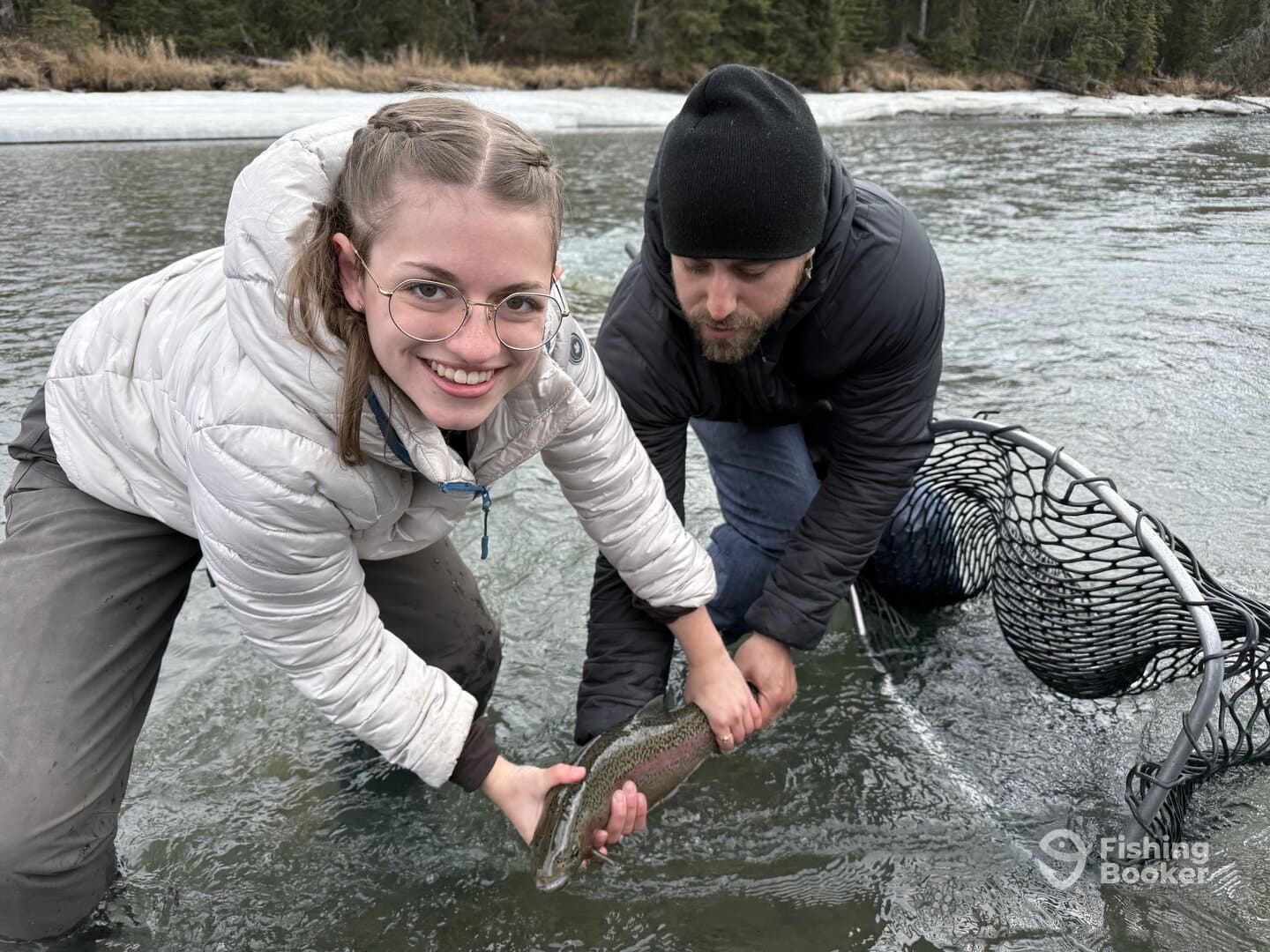 A young angler and an adult are joyfully holding a Rainbow Trout in a river setting, showcasing a successful catch during a fishing trip.
