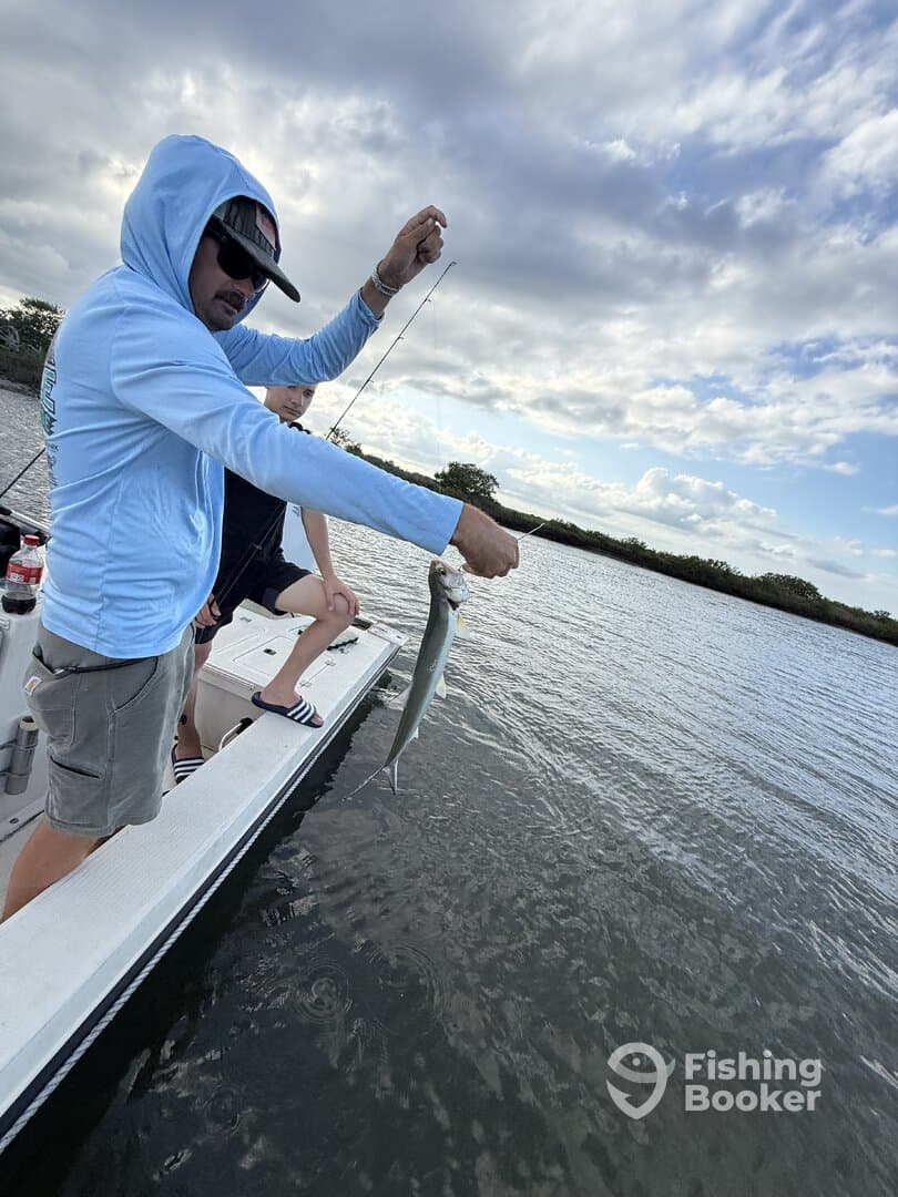Angler reeling in a fish while fishing from a boat in a scenic waterway.